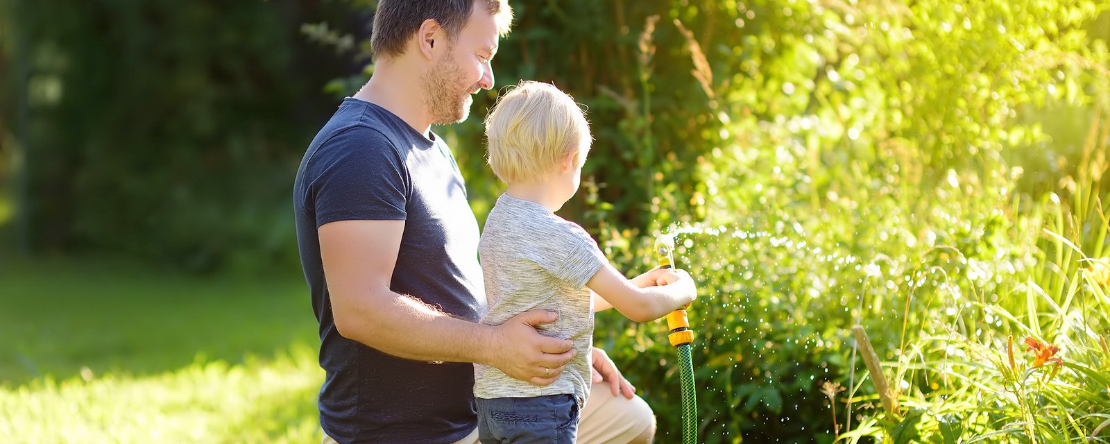 Funny little boy with his father playing with garden hose in sunny backyard. Preschooler child having fun with spray of 